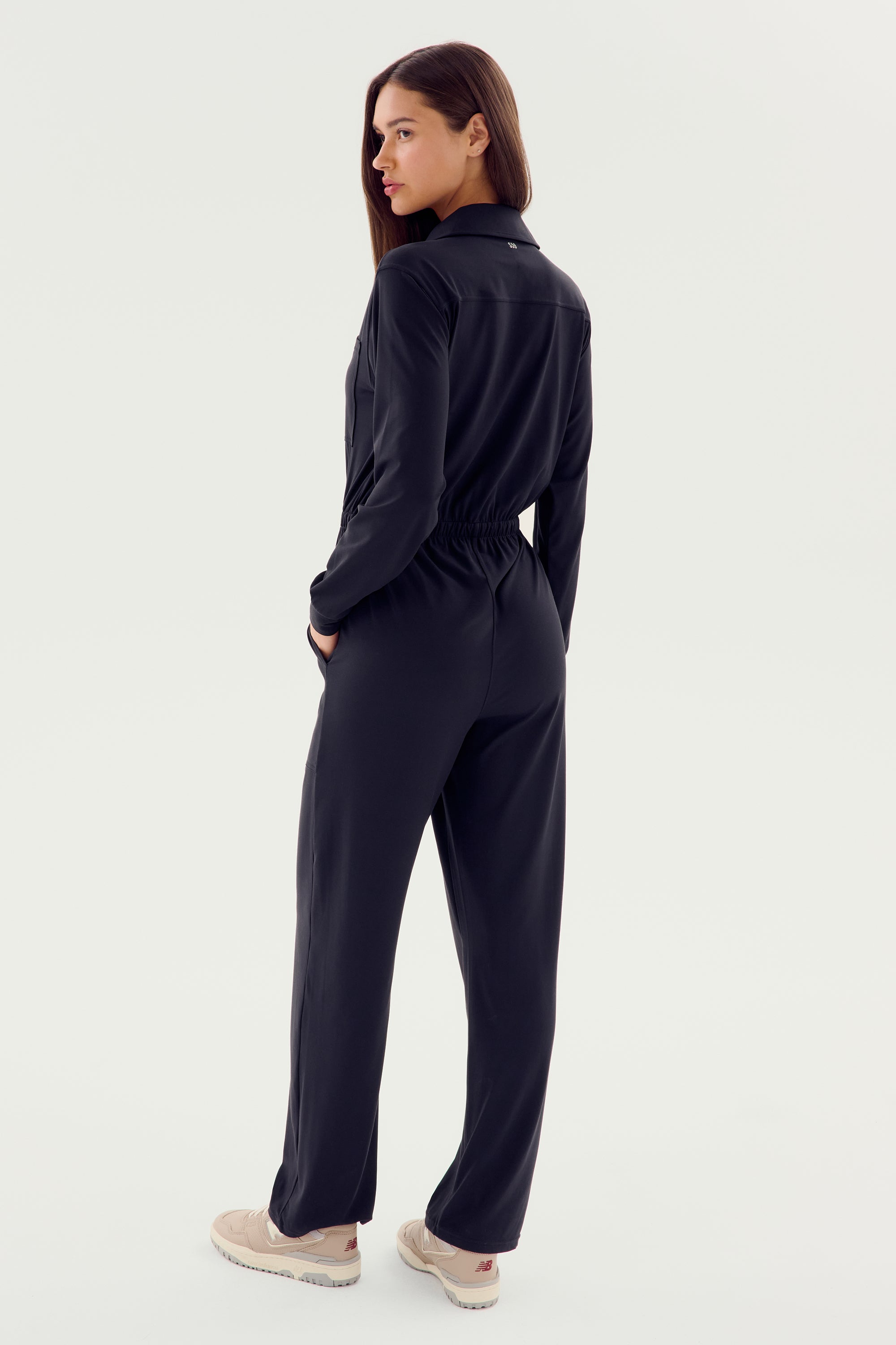 A person with long hair exudes modern style in a SPLITS59 Ollie Airweight Boiler Suit - Black, complemented by beige sneakers. With one hand in their pocket, they confidently look over their shoulder.
