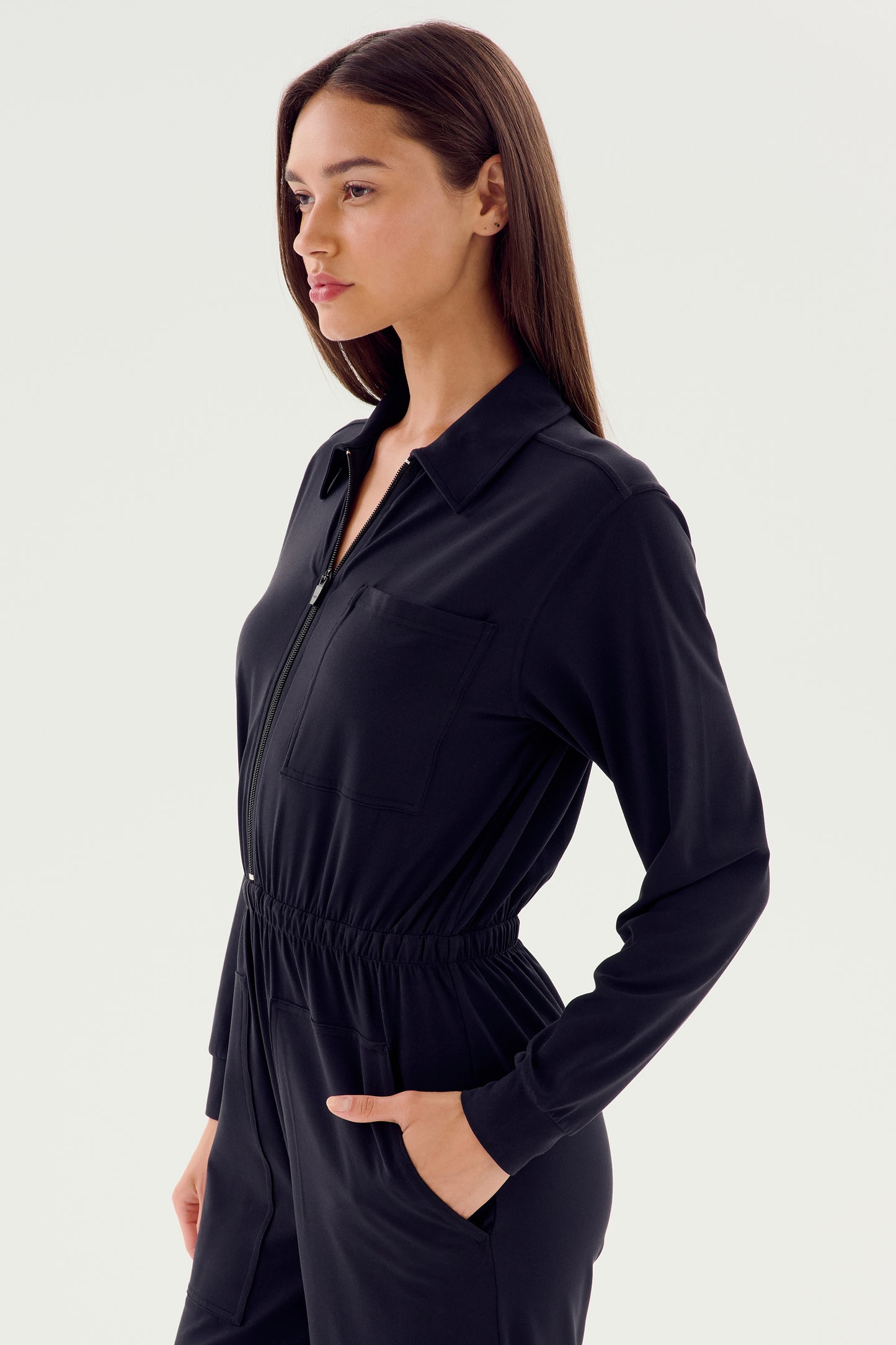A person in a black, long-sleeve Ollie Airweight Boiler Suit by SPLITS59 stands with hands in pockets, showcasing modern style against a plain background while gazing to the side.