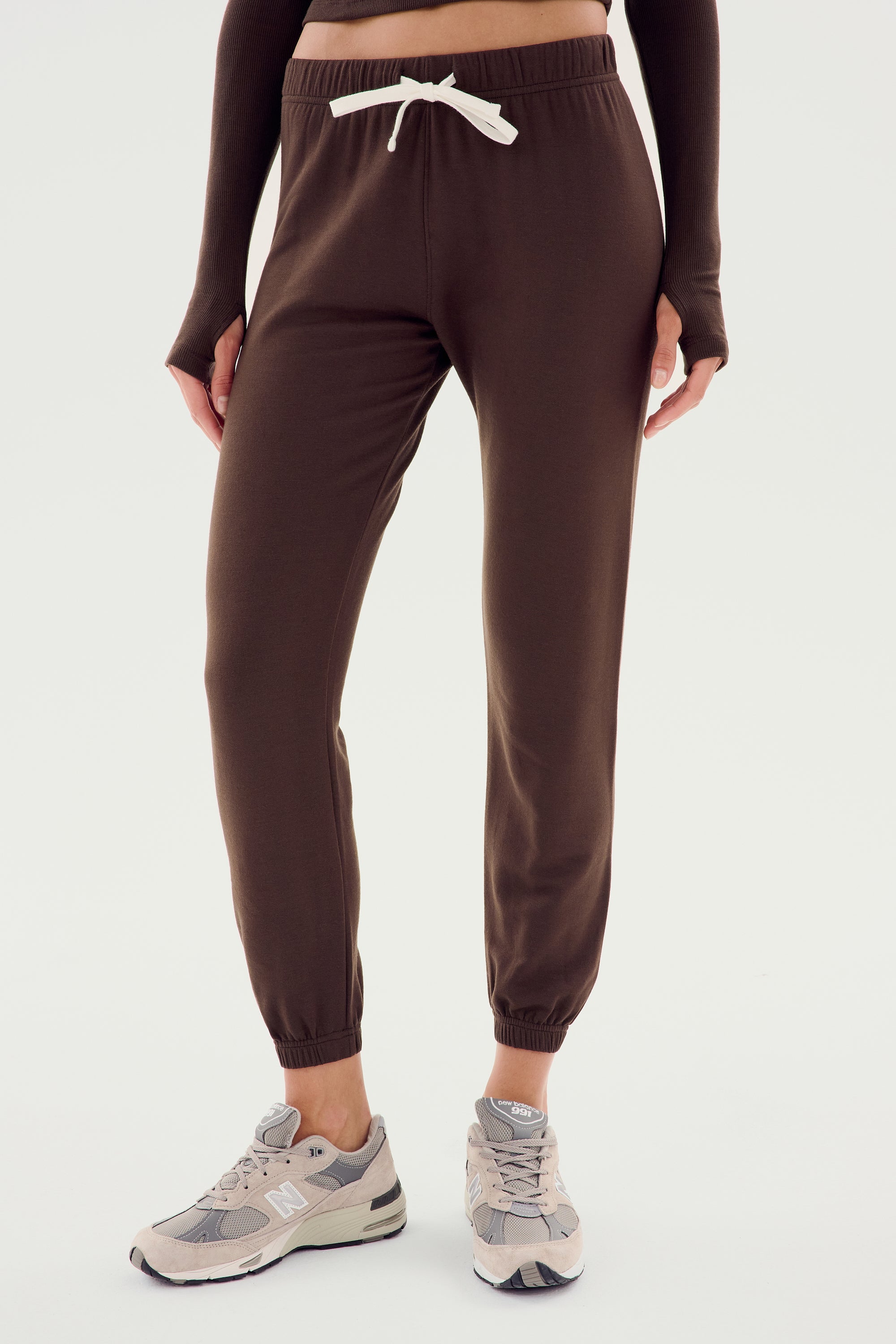 A person is wearing the Sonja Fleece Sweatpant - Dark Chocolate by SPLITS59, featuring a white drawstring, paired with beige sneakers. Perfect for cool weather workouts, the minimalistic backdrop is plain and white.